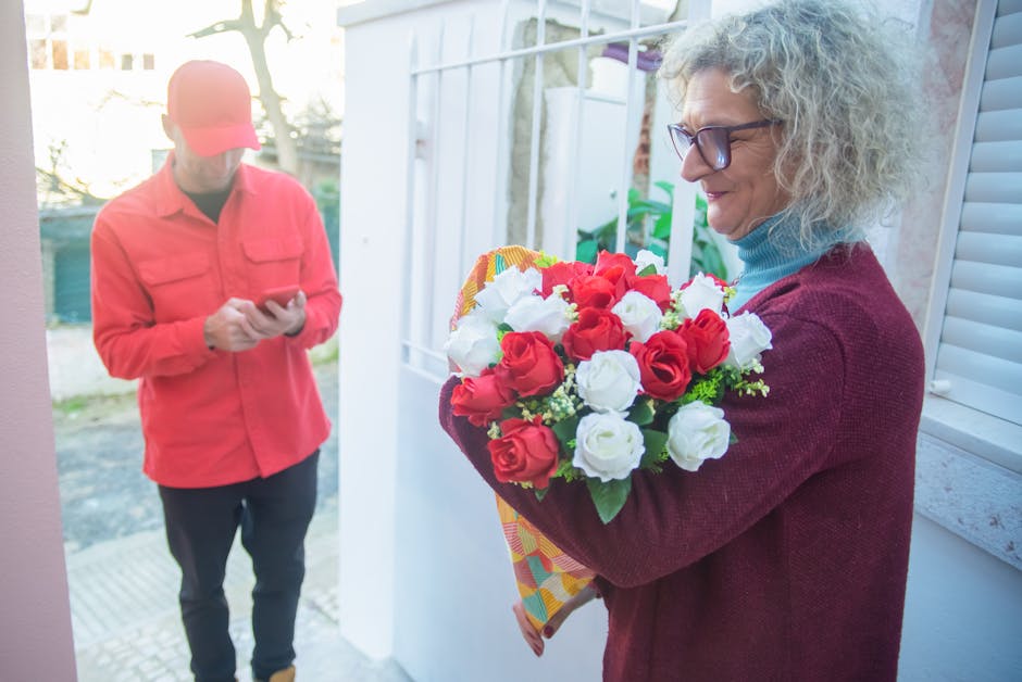 A woman with curly gray hair, wearing glasses and a burgundy sweater over a light blue turtleneck, holding a vibrant bouquet of fresh red and white roses wrapped in colorful paper. She is standing nea