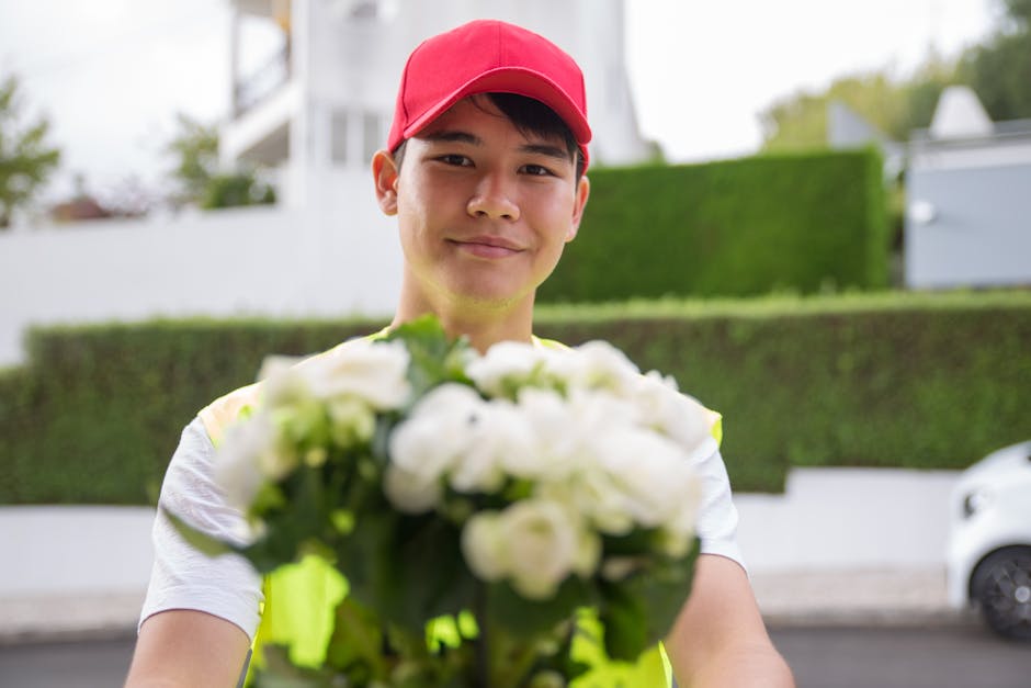 A delivery person wearing a red cap and a high-visibility vest is smiling and offering a bouquet of fresh white flowers with green foliage. The bouquet is wrapped in clear cellophane, and the flowers