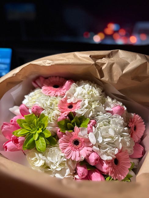 A bouquet of fresh flowers contains white hydrangeas and pink gerbera daisy flowers, arranged with bright green foliage, wrapped in light brown kraft paper, and presented on a surface with a blurred c