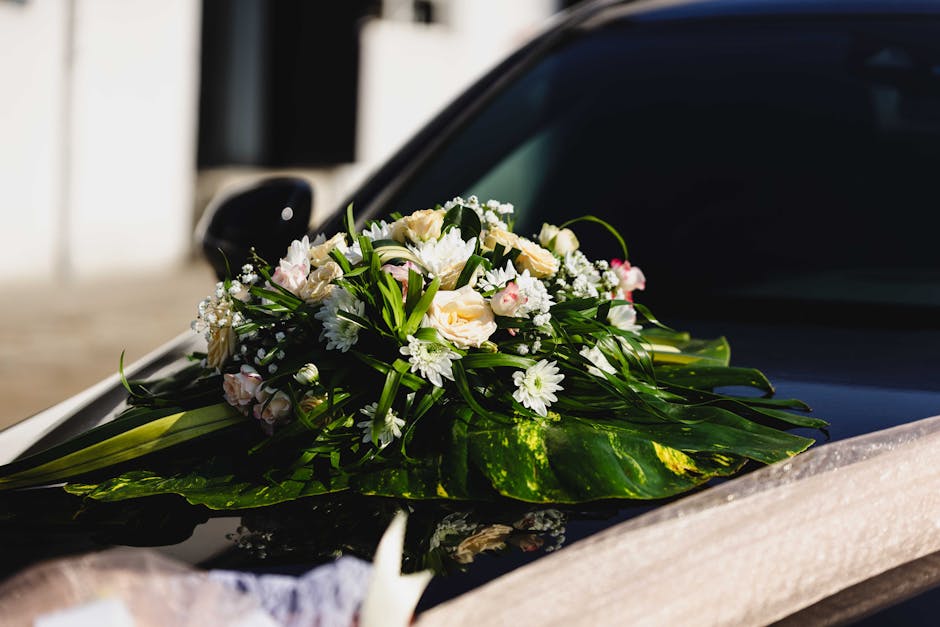 A beautifully crafted bouquet of fresh white and light pink roses, daisies, and small filler flowers, arranged with lush green foliage and long leaves, resting on the glossy black hood of a car. The b