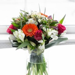 This stunning bouquet captures the essence of floral artistry, thoughtfully composed by Stanmore's experienced florists to bring colour, structure, and warmth into any space. Creamy peach gerbera daisies fan out, their petals softly framing bold, festive orange centres that seem to twinkle in the day's gentle light, echoing the vibrancy of local Stanmore celebrations. The depth of fuchsia roses adds drama and refinement to the composition, while ivory alstroemeria and dainty white fillers create luminous, garden-fresh highlights. Broad, deep green aspidistra leaves gently wrap around the bouquet's base, suggesting the embrace of Stanmore's leafy streets and tranquil parks. Slender, pointed greens and textural sprigs are nestled carefully amongst the flowers, heightening contrast and fullness. All is presented in a tall, slender glass vase, the clear surface showcasing the natural tangle of stems, furthering the bouquet's sense of freshly gathered abundance. Filtered daylight enhances the delicate interplay of hues and forms, imbuing the bouquet with a serene, joyful presence-perfect for a brunch, heartfelt gathering, or marking the beginning of a new chapter. Every stem reflects not only the florist's craft but the welcoming character and green spaces of Stanmore.